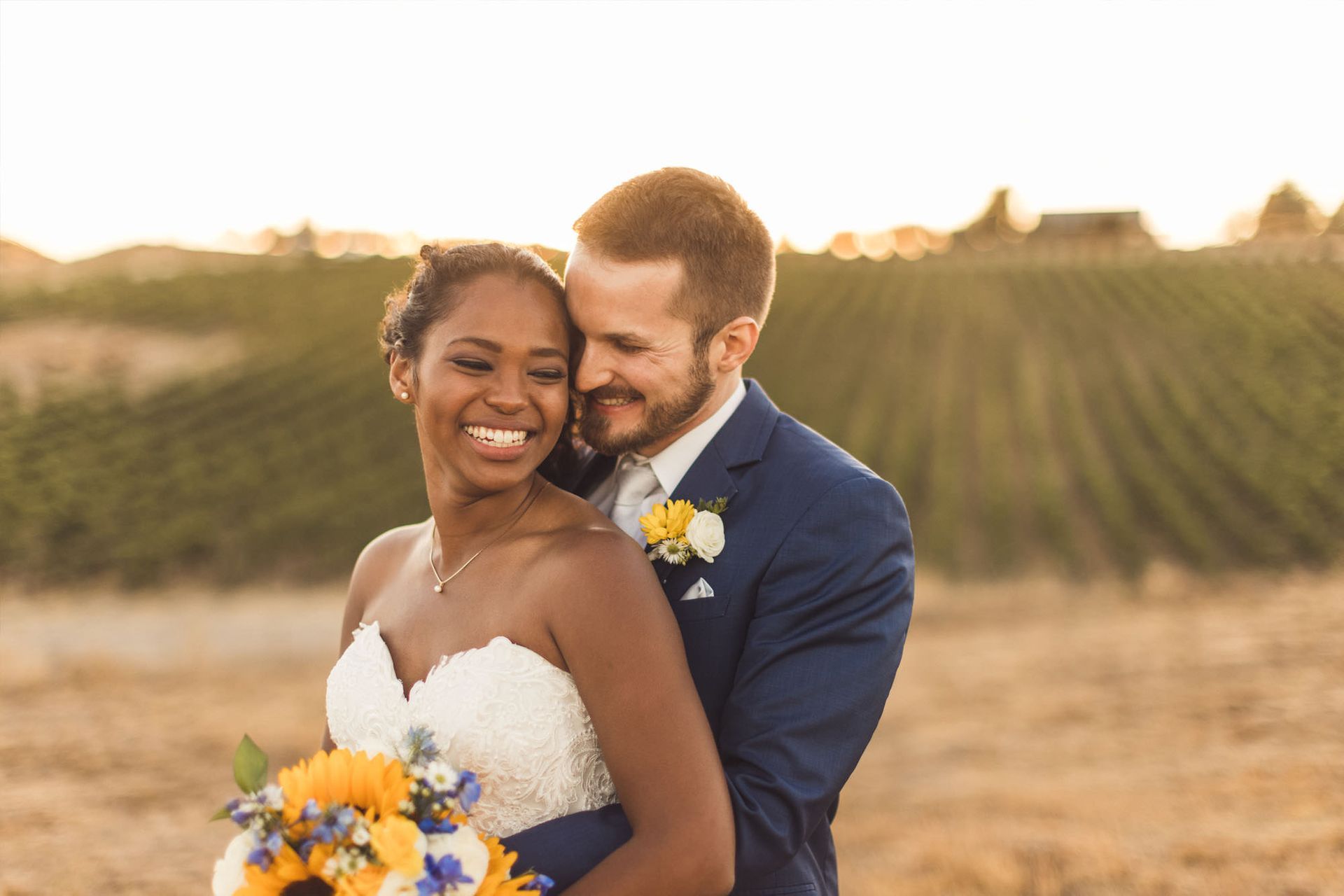 Bride and groom embracing, smiling, and looking at each other. They are standing in front of a vineyard at sunset.