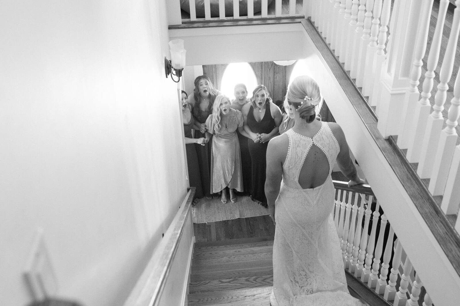Bride descends stairs; bridesmaids react with surprise. Black and white photo.