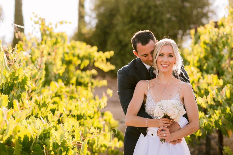 Wedding couple embraces in a vineyard. The woman holds a bouquet, the man kisses her neck; golden sunlight.