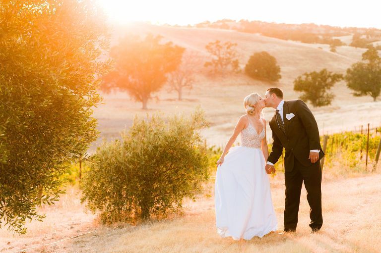Couple kissing, holding hands, in wedding attire, sunset over rolling hills, vineyard.