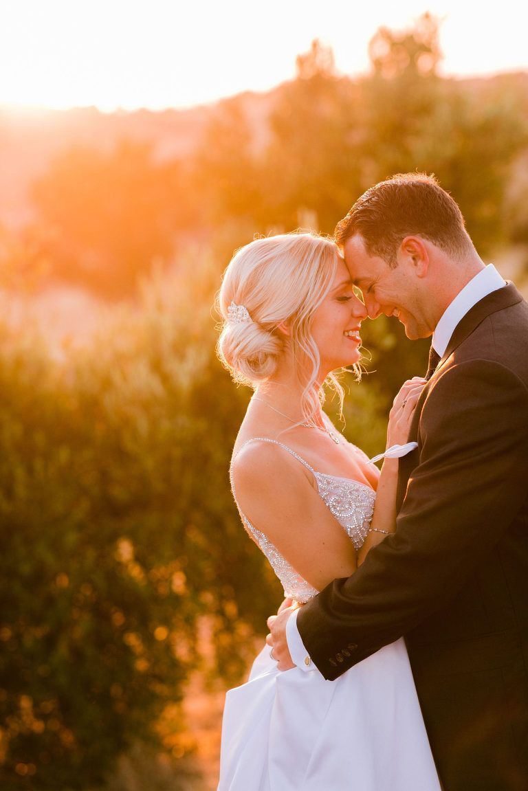 Bride and groom embrace at sunset, backs to the camera, smiling, warm light, outdoor setting.