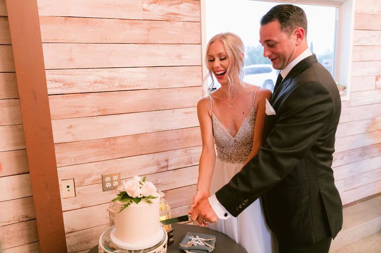 Couple cuts wedding cake; woman laughs, man smiles; indoors, light wood paneling.