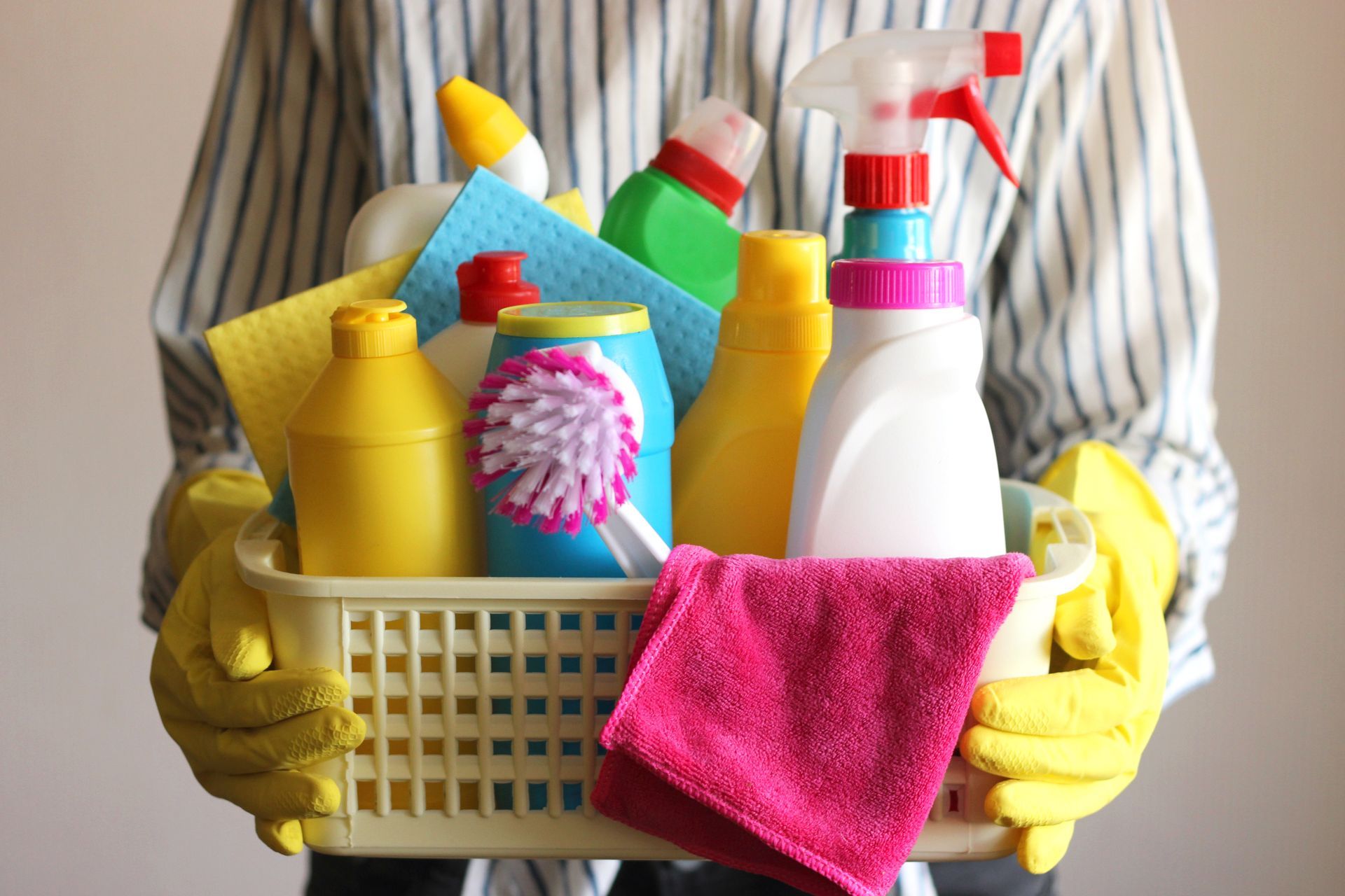 Person holding a basket of colorful cleaning supplies and a pink cloth. Person holding a basket of colorful cleaning supplies and a pink cloth.