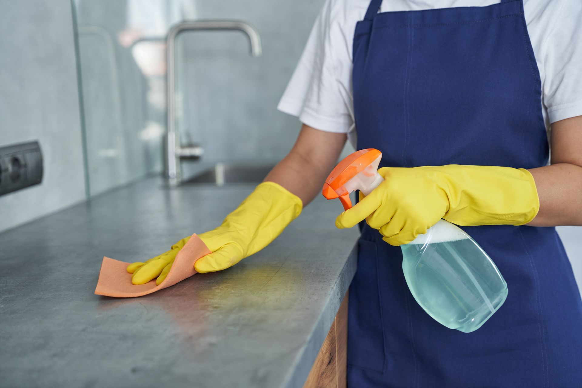Person cleaning a kitchen counter with a cloth and spray bottle, wearing yellow gloves and a blue apron Person cleaning a kitchen counter with a cloth and spray bottle, wearing yellow gloves and a blue apron
