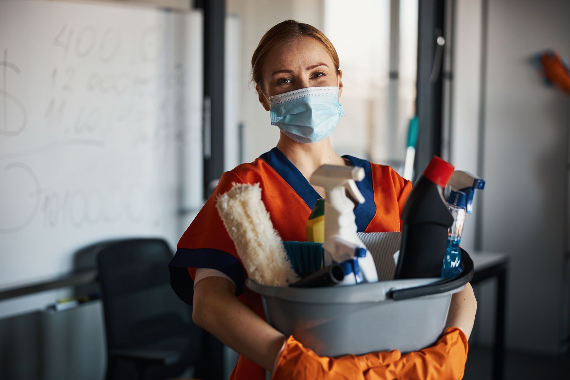 Cleaner in mask holding cleaning supplies in a classroom Cleaner in mask holding cleaning supplies in a classroom