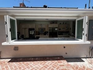 Open white kitchen pass-through window onto a bright tiled patio, with counter and shelves visible inside.