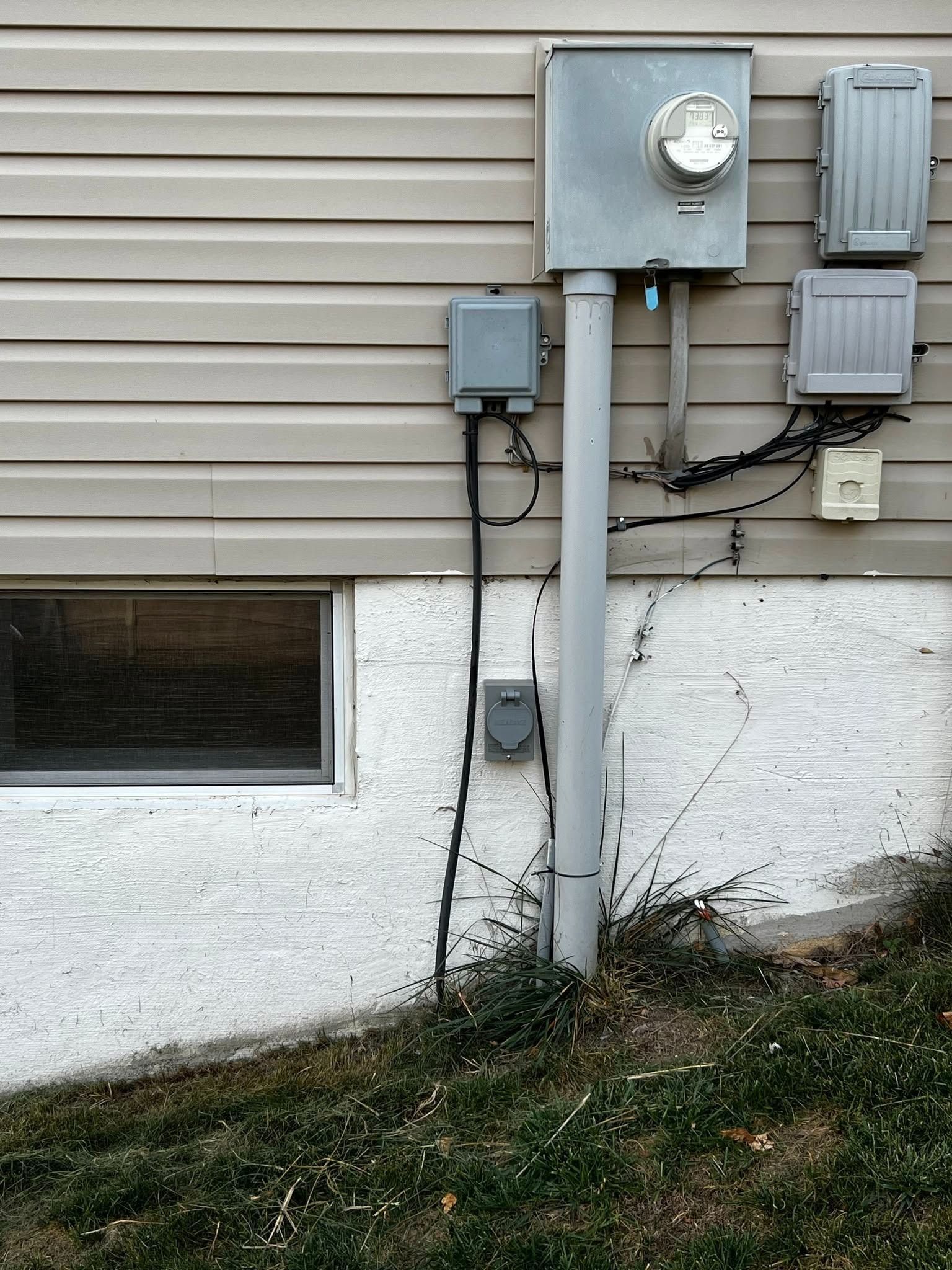 Electric meter and utility boxes mounted on a house exterior with siding and foundation wall.