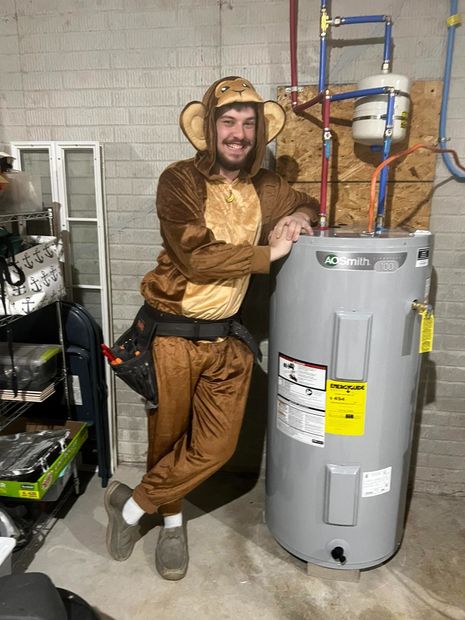 Person in a monkey costume posing beside a water heater in a basement utility room.
