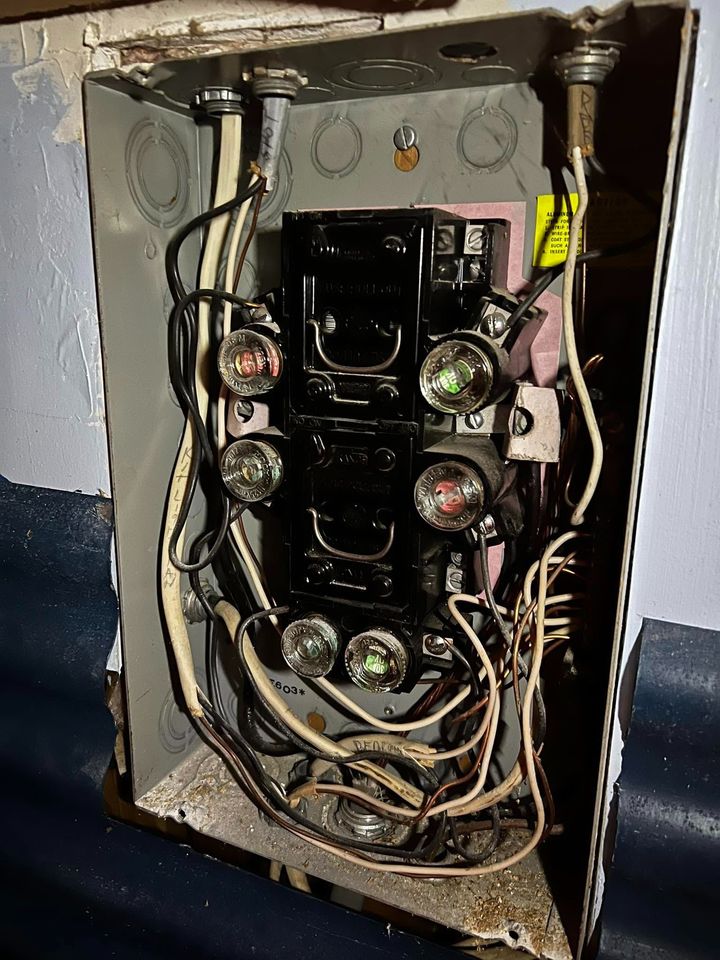 Open electrical panel with tangled wires and circuit breakers exposed inside a wall.