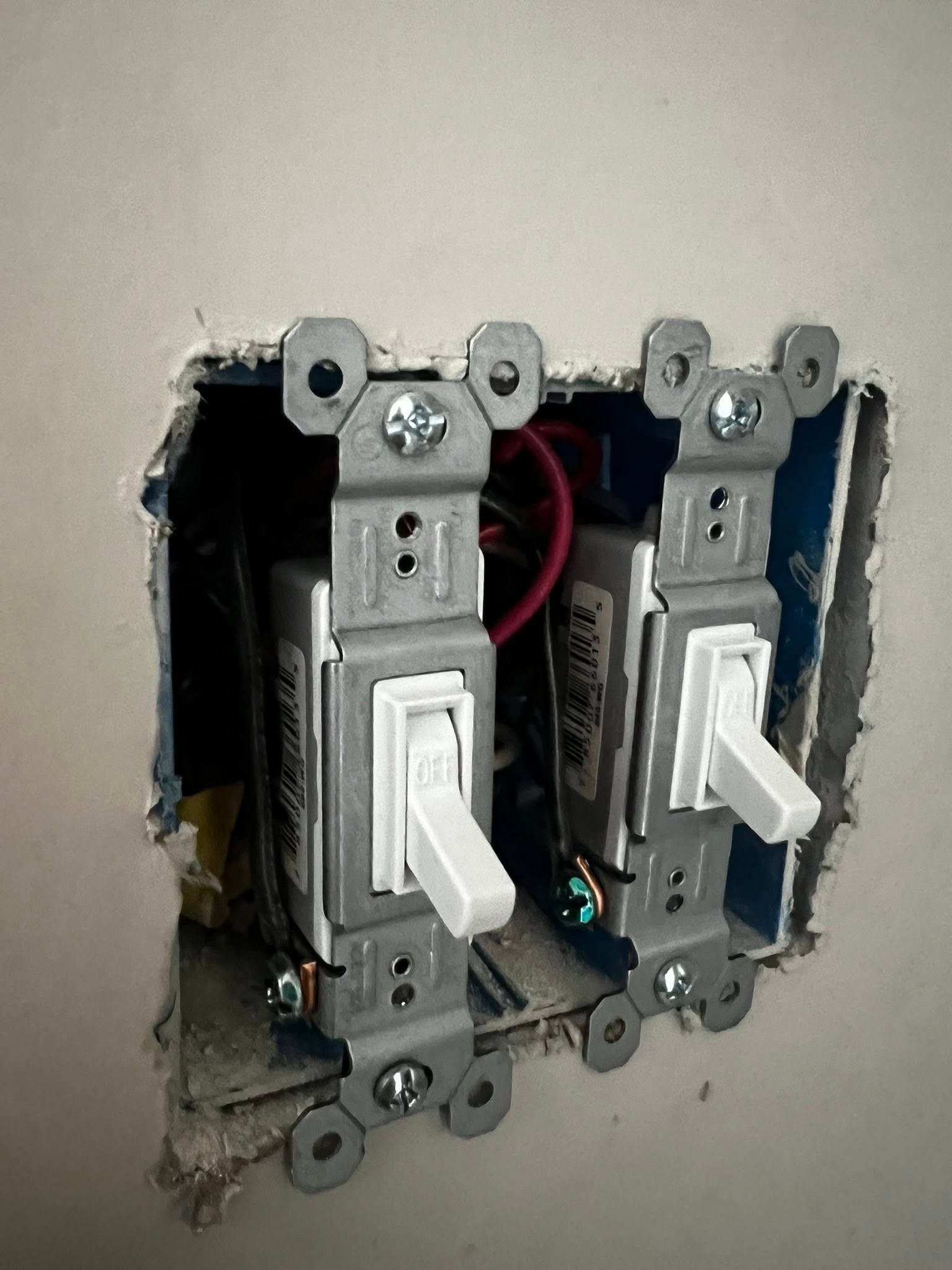 Two white toggle light switches in a wall opening, with exposed wiring and metal mounting brackets