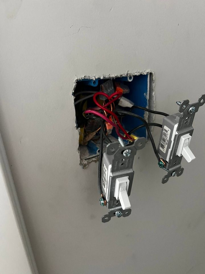 Open electrical outlet box with exposed wires and two wall switches on a gray wall