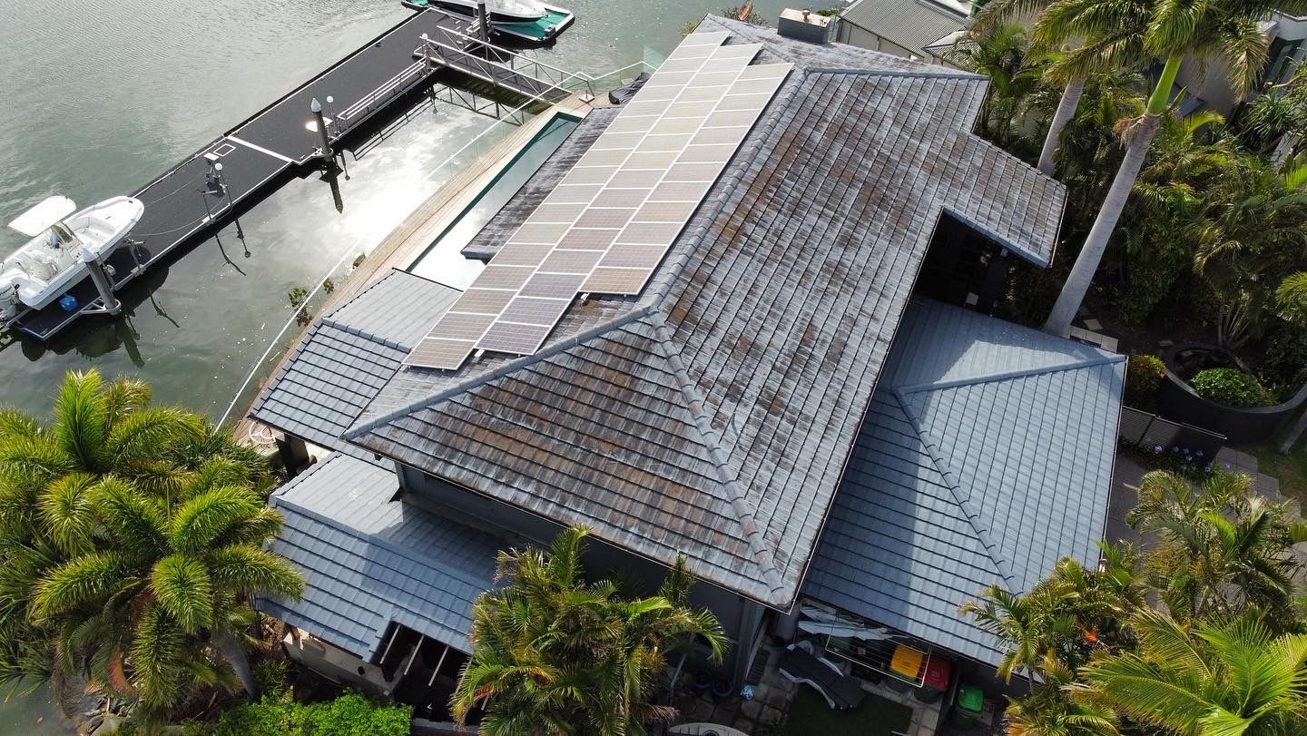 Aerial view of a house with solar panels on a gray tiled roof, next to a dock with boats and water. - Roof Painting in Gold Coast,QLD