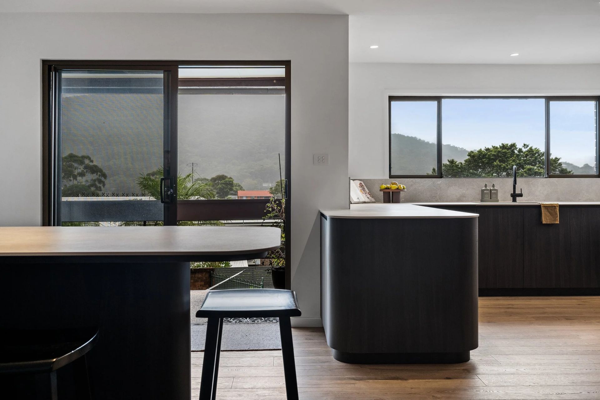 Modern kitchen with dark cabinetry, white countertops, wooden floors, large windows — Onyx Detailed Joinery In Unanderra, NSW