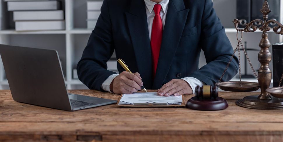Businessperson in suit writing at a desk with laptop, papers, gavel, and scales of justice