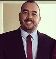 Um homem com barba está vestindo terno e gravata e sorrindo.
