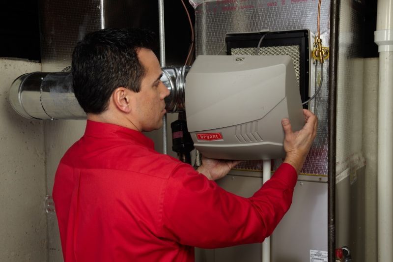 Man in red shirt installing a humidifier on a furnace in a utility room.