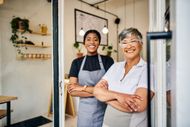 Two smiling people stand in a doorway of a bright shop, one in an apron, arms crossed.