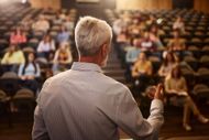 Speaker addressing a seated audience in a lecture hall, seen from behind with warm lighting.