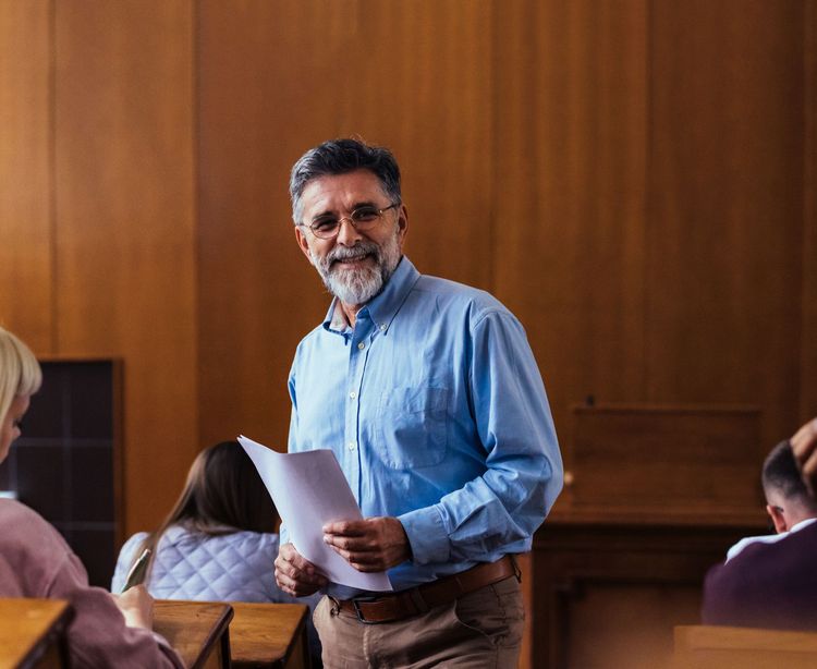 Man in blue shirt holding papers in a wood-paneled meeting room, standing among seated attendees