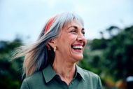 Smiling person with gray hair in a green shirt outdoors with blurred trees behind them