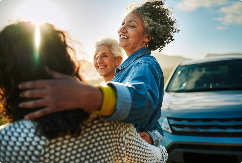 Three people smiling and hugging outdoors by a vehicle in bright sunlight