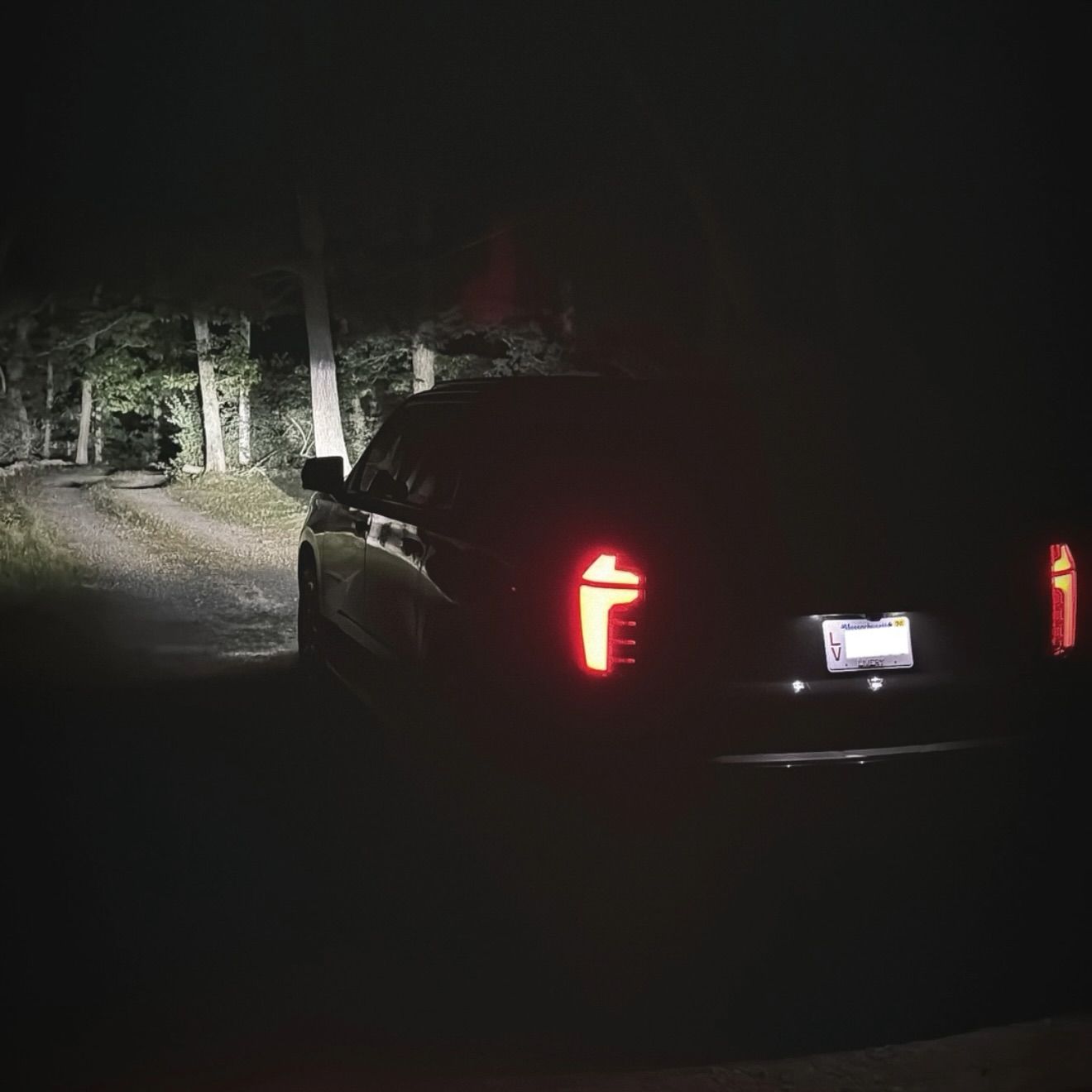 Black SUV parked on a dark road at night, headlights illuminating a stone wall and trees.