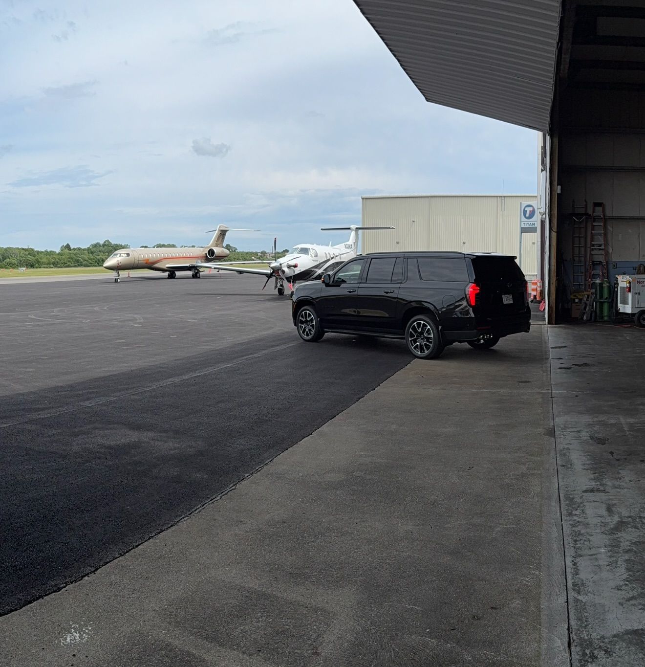 Airport tarmac with a black SUV parked near a hangar and a plane in the background