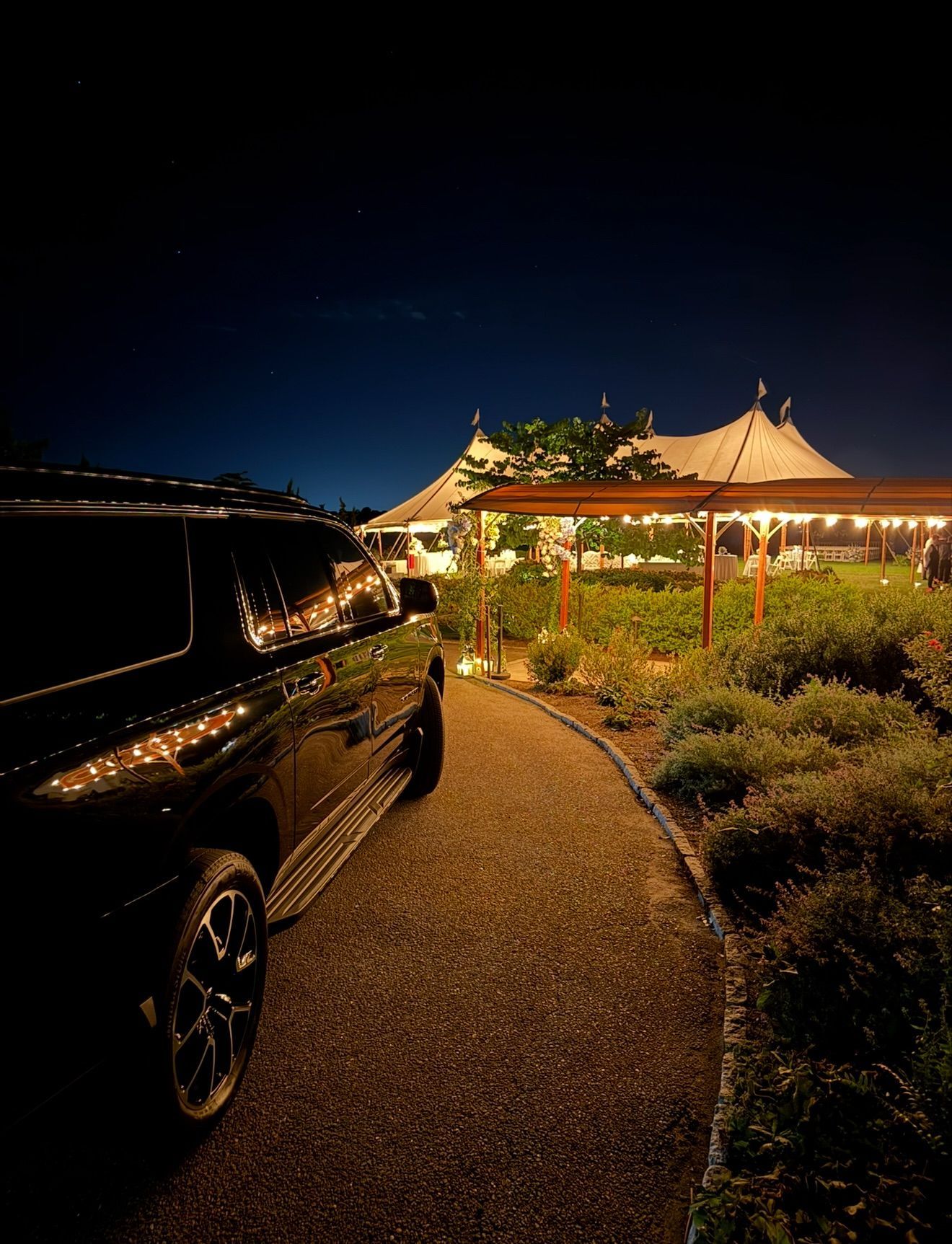 Black car beside a lit garden pavilion at night, with a curved path and dark sky overhead