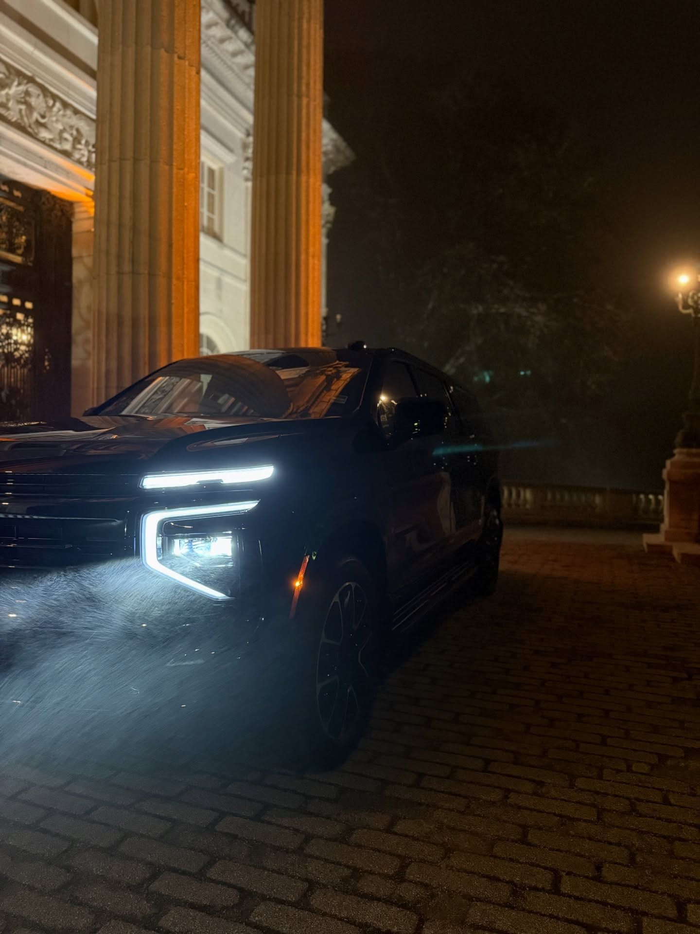 Dark SUV with bright headlights parked at night beside a lit building with columns.