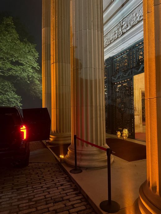 Nighttime entrance with tall lit columns, red rope barriers, and a black SUV at the curb.
