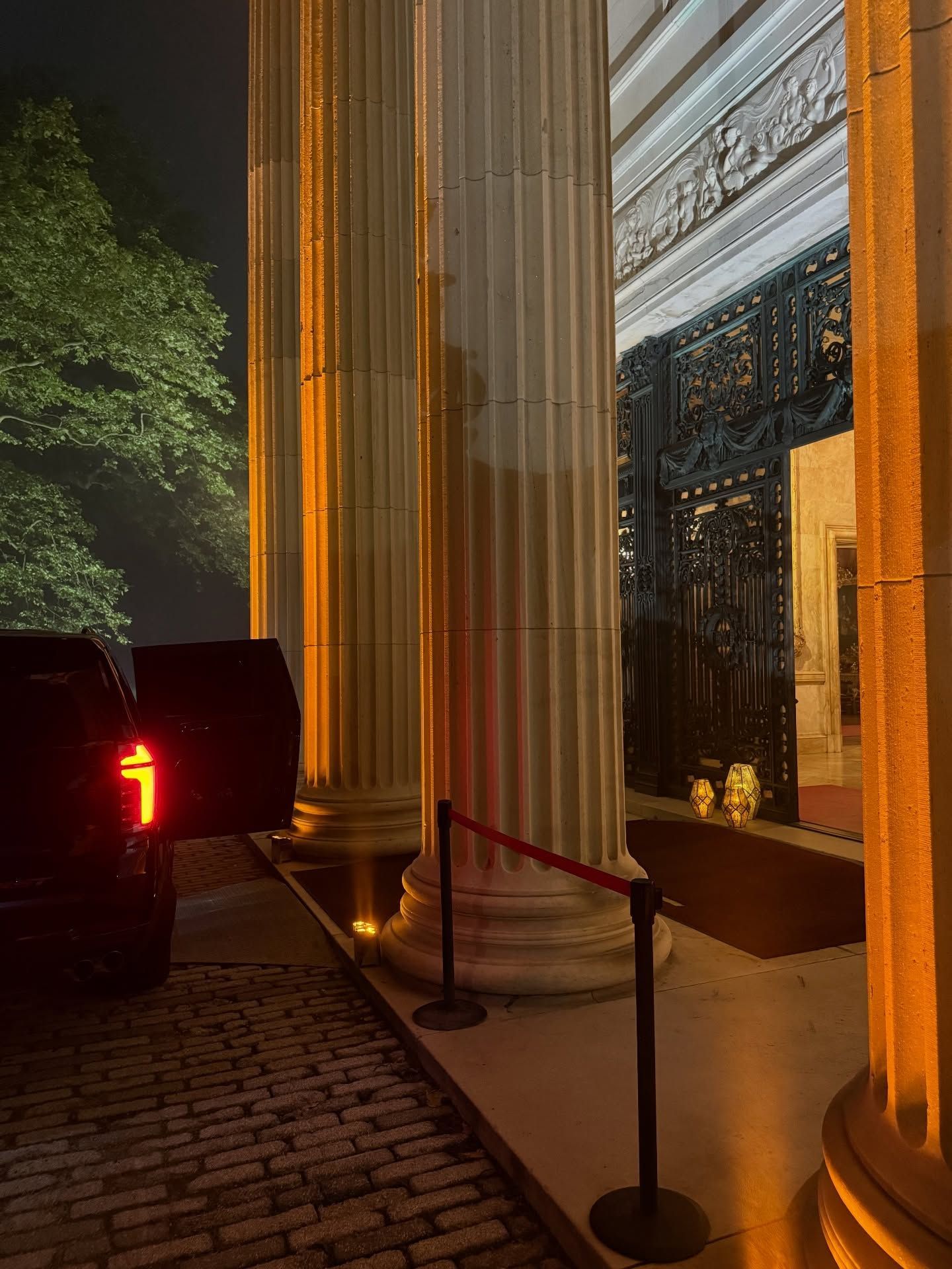 Lit columns and ornate building entrance at night, with a black SUV parked beside a cobblestone walkway.