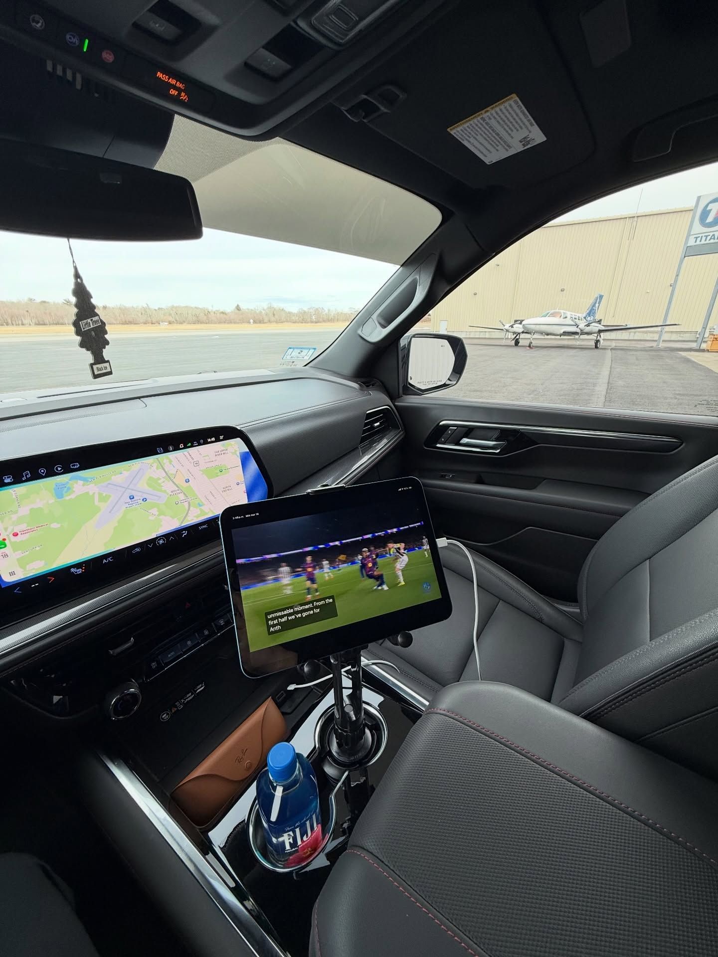 Car interior with two displays; map on dashboard and soccer match on center screen, parked by water.