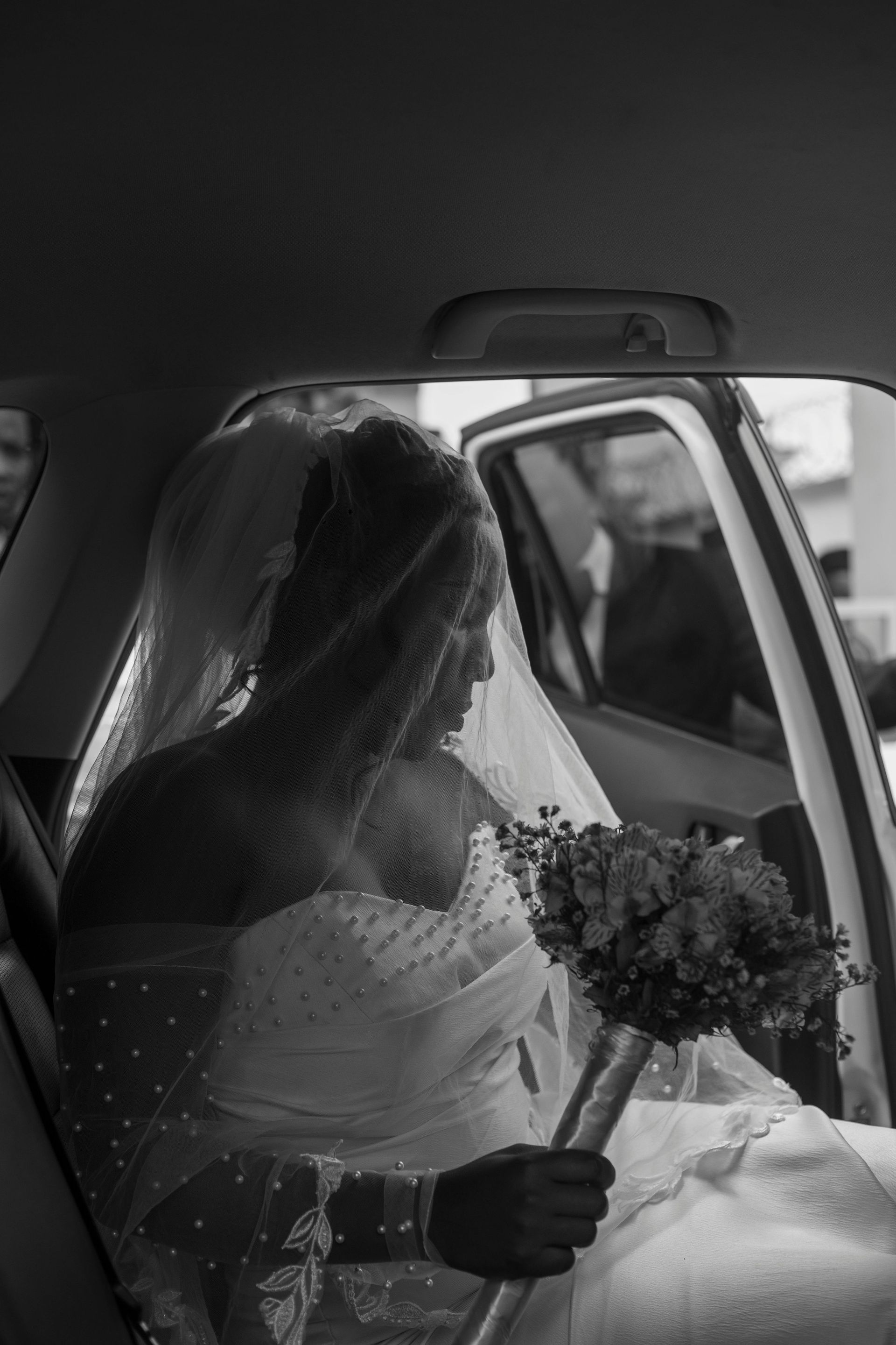 Bride in a wedding dress and veil sits in a car holding a bouquet.