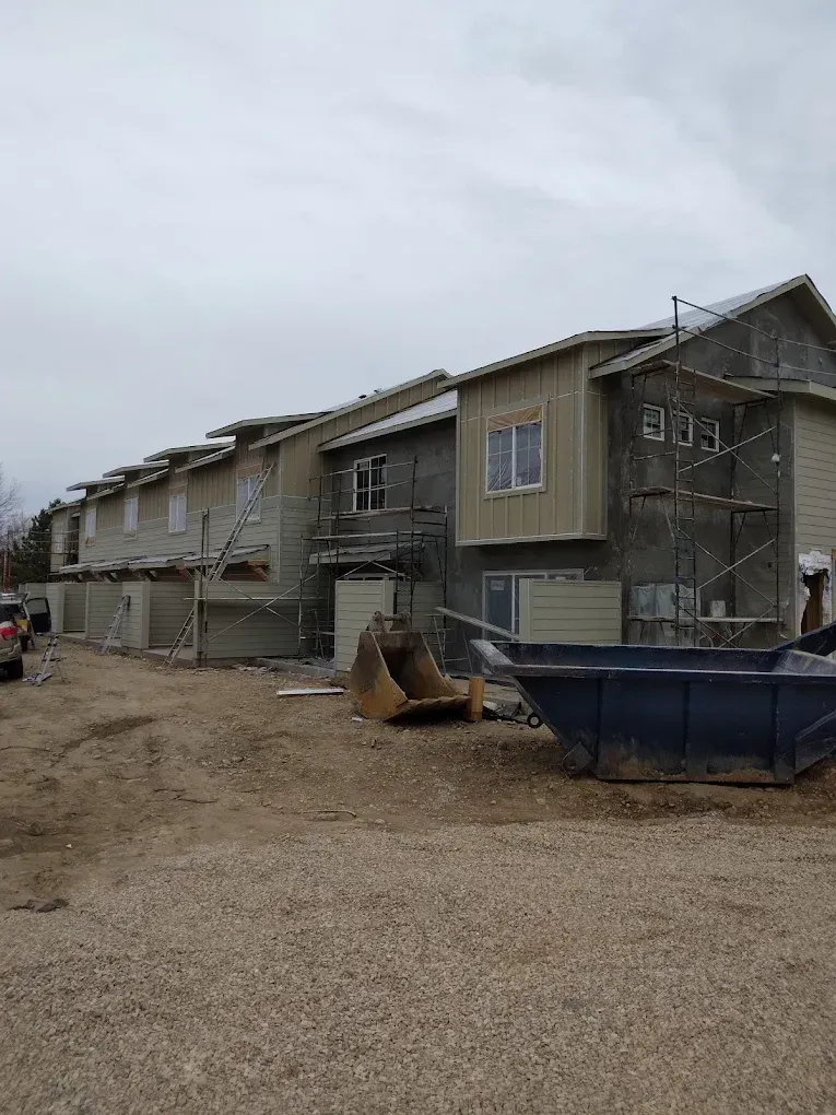 Partially built apartment row with scaffolding, dirt lot, and construction dumpster under overcast sky
