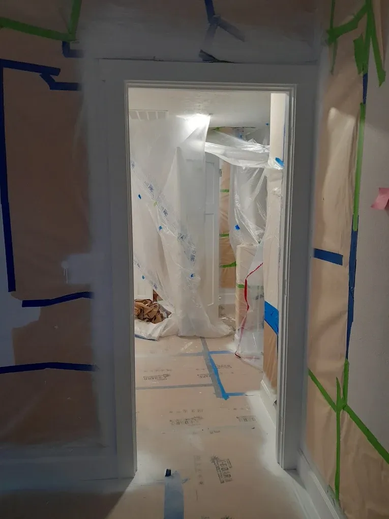 Plastic-covered hallway with blue painter’s tape and a doorway leading into a renovated room.