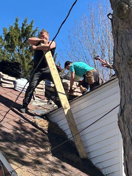 Two workers on a roof installing a long board near a white siding wall under a clear blue sky
