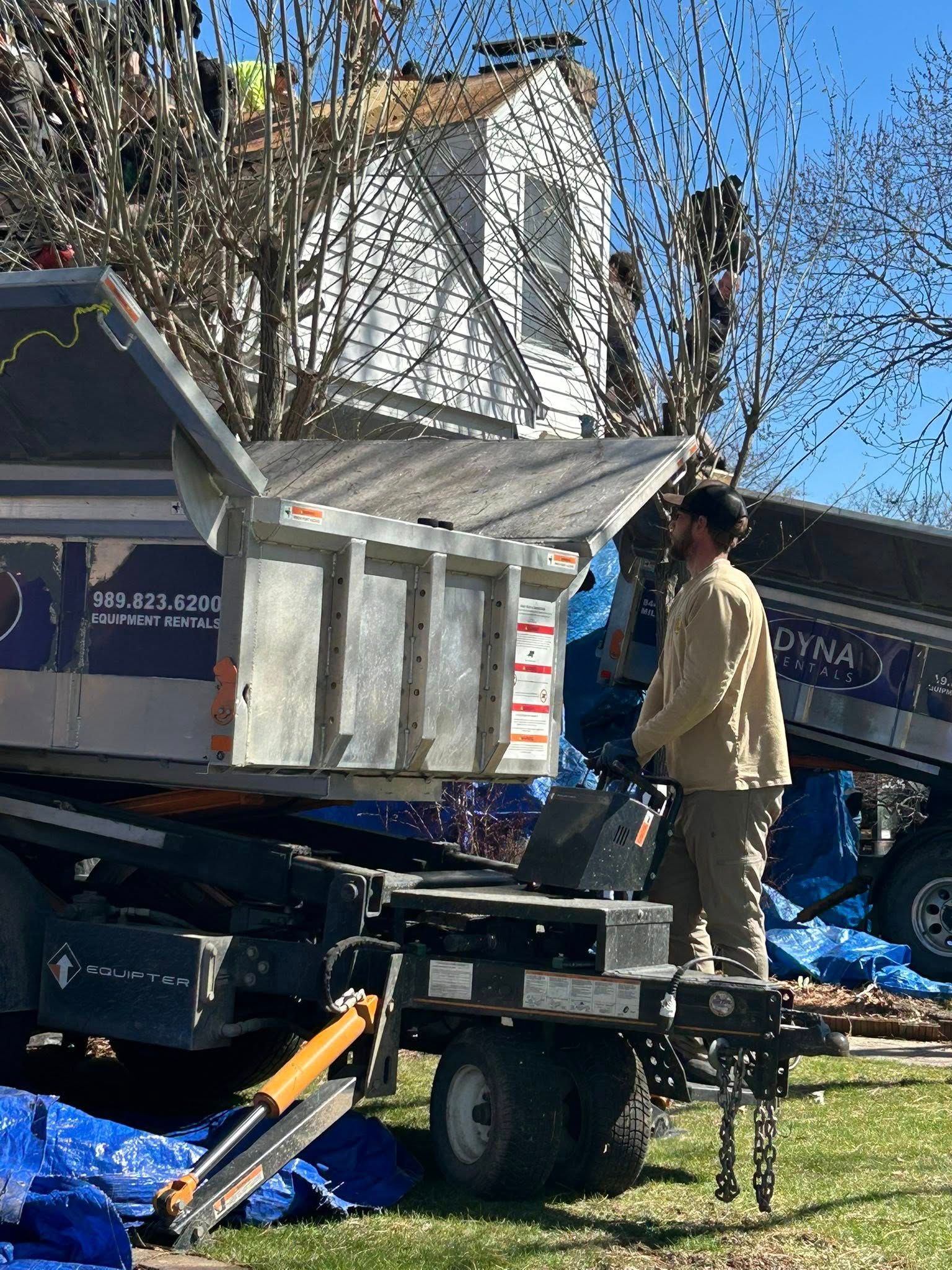 Worker loading a house model onto a trailer in a yard with blue tarps and trees