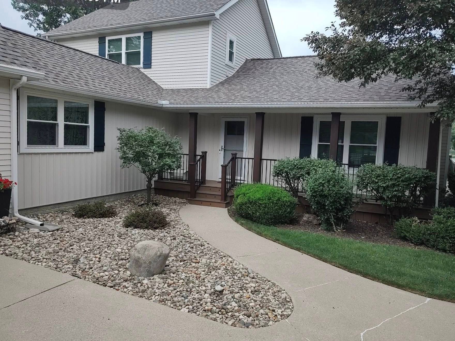 Suburban house entrance with curved concrete walkway, white siding, stone landscaping, and green shrubs