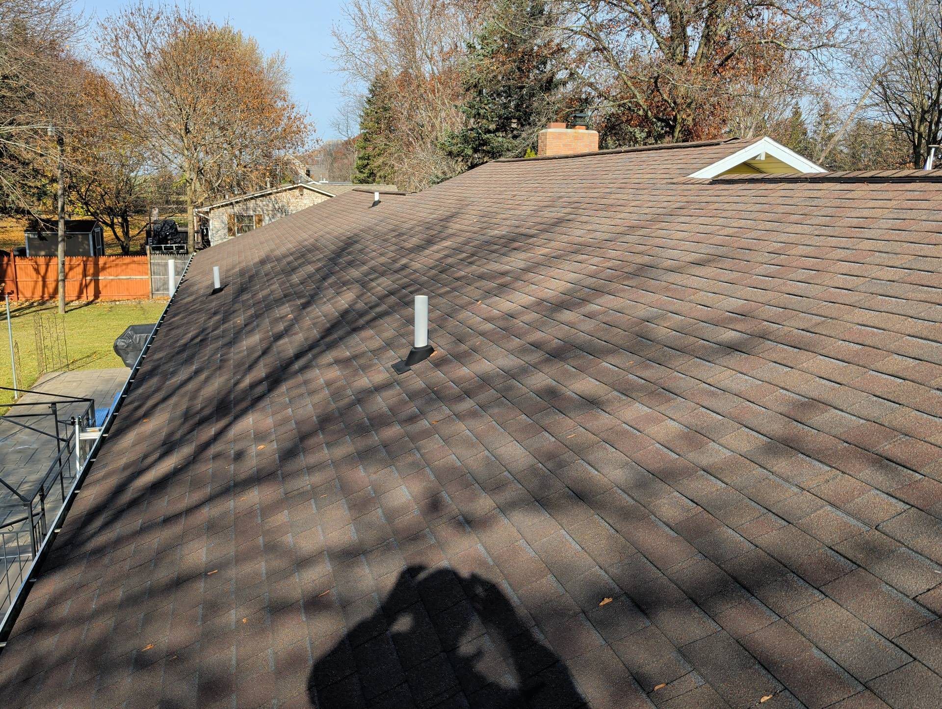 Sloped brown shingle roof with a shadow, against trees and neighboring houses on a sunny day