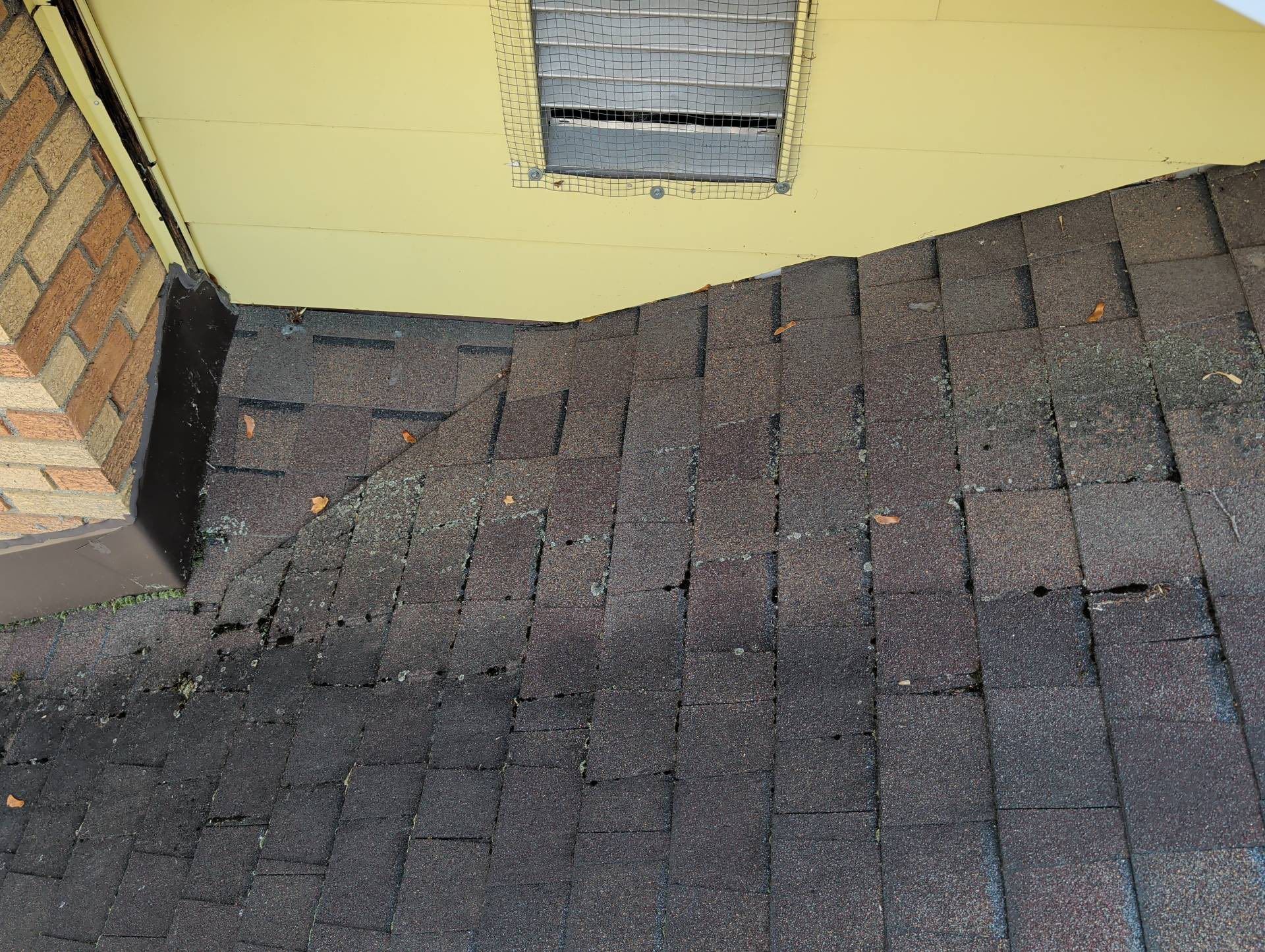 Shingled roof with a damaged area near a brick wall and yellow exterior siding