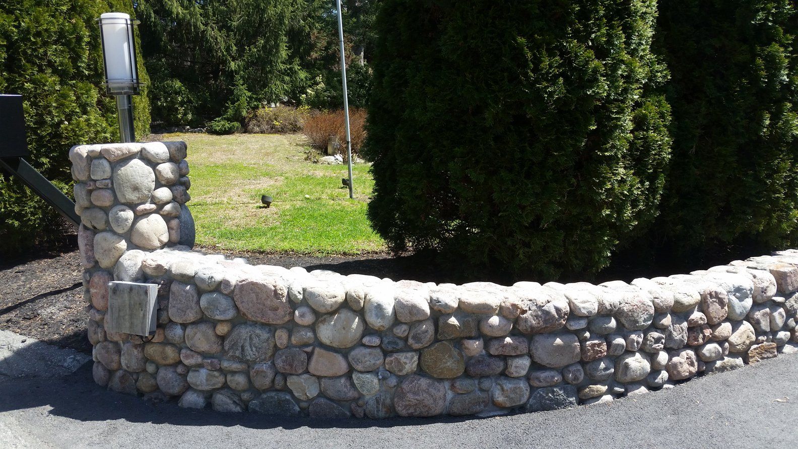 Stone wall and pillar on edge of driveway, with grassy area and green shrubs in the background.