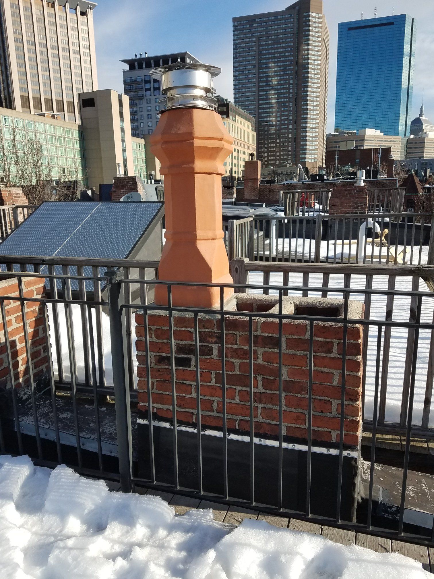 Red brick chimney on a rooftop with snow, black railing, city buildings in the background.