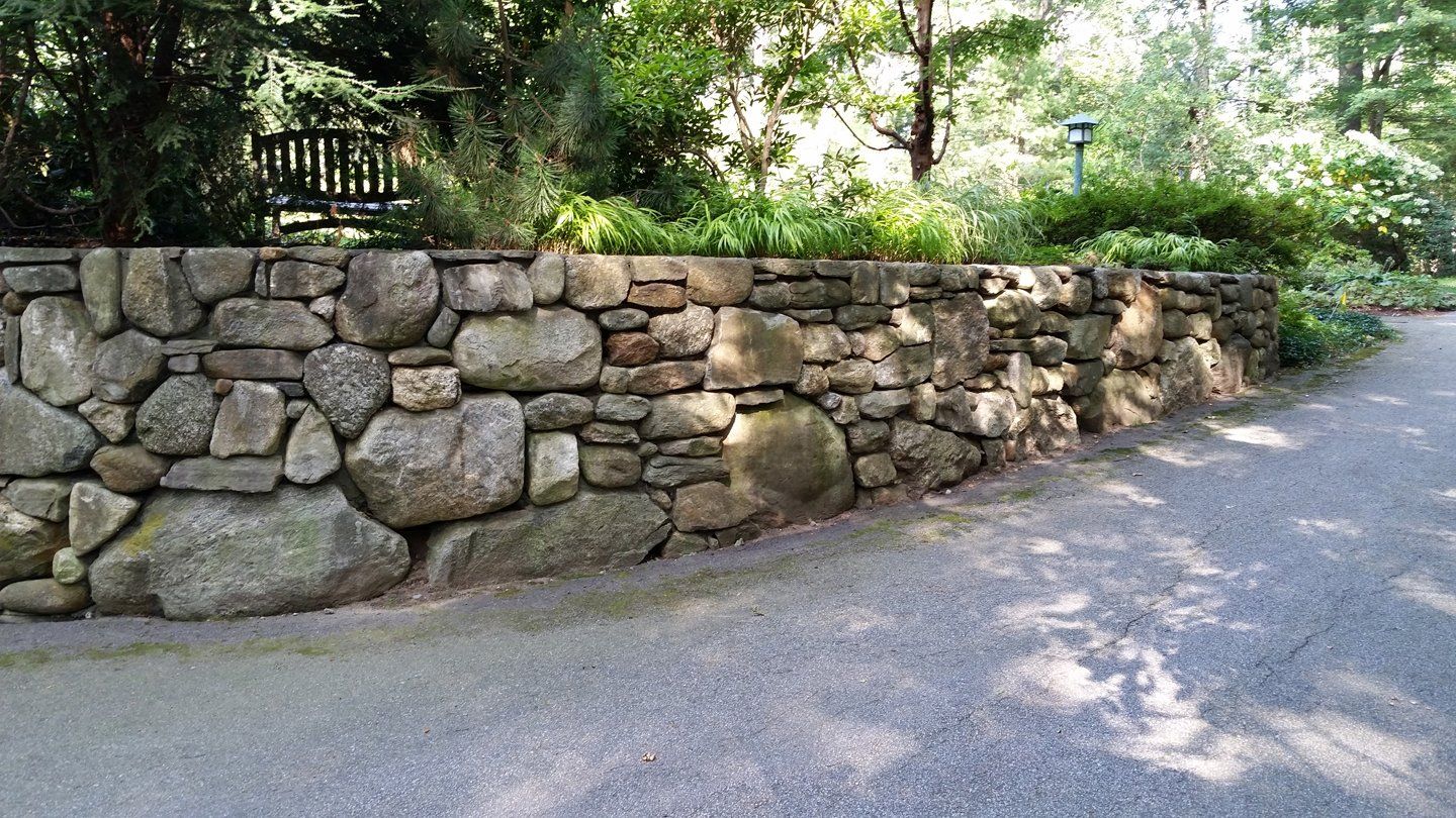 Stone wall bordering a gravel path, with greenery and trees in the background.
