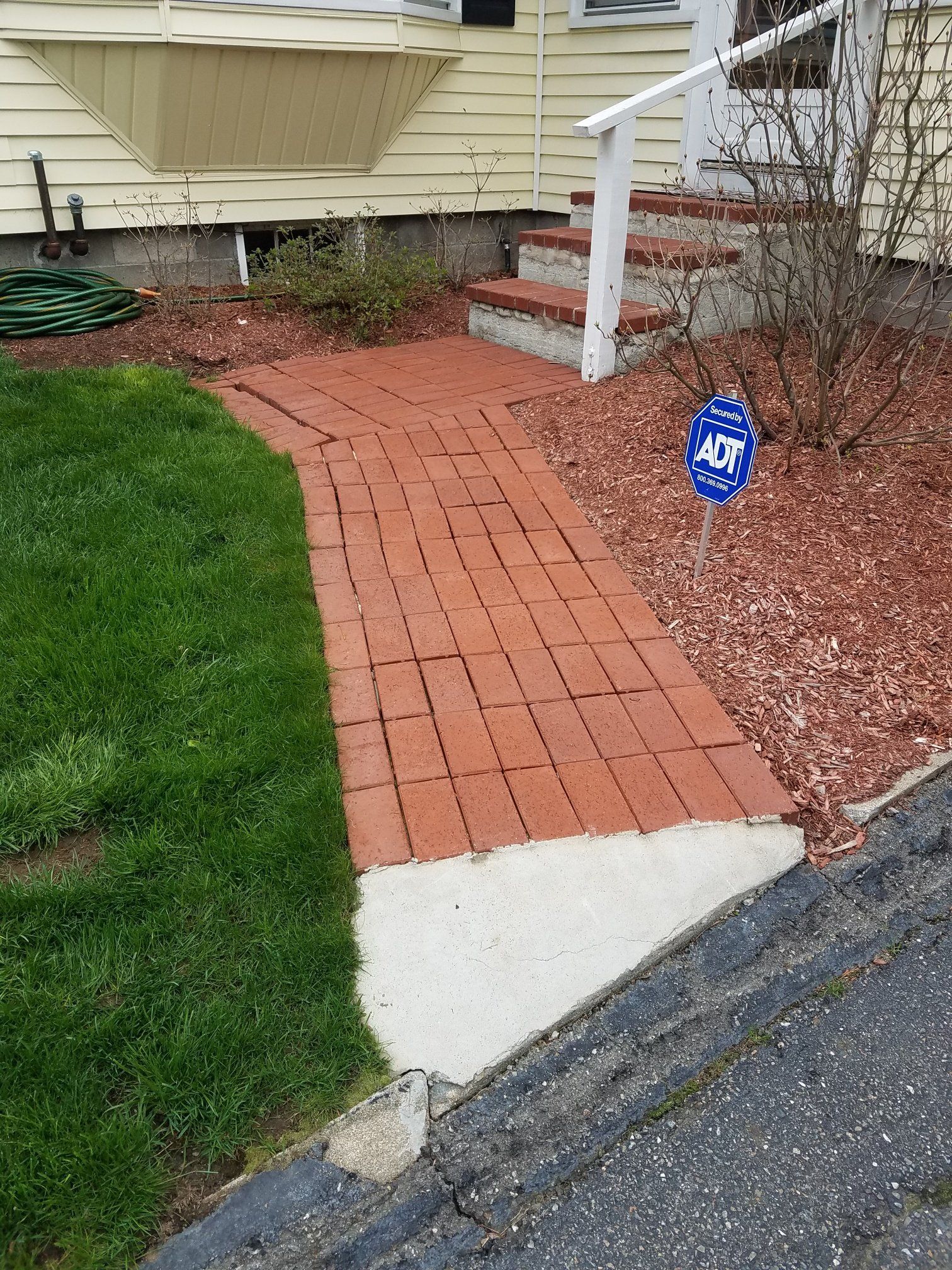 Brick walkway leading to house entrance with steps, bordered by grass and mulch, concrete curb in foreground.