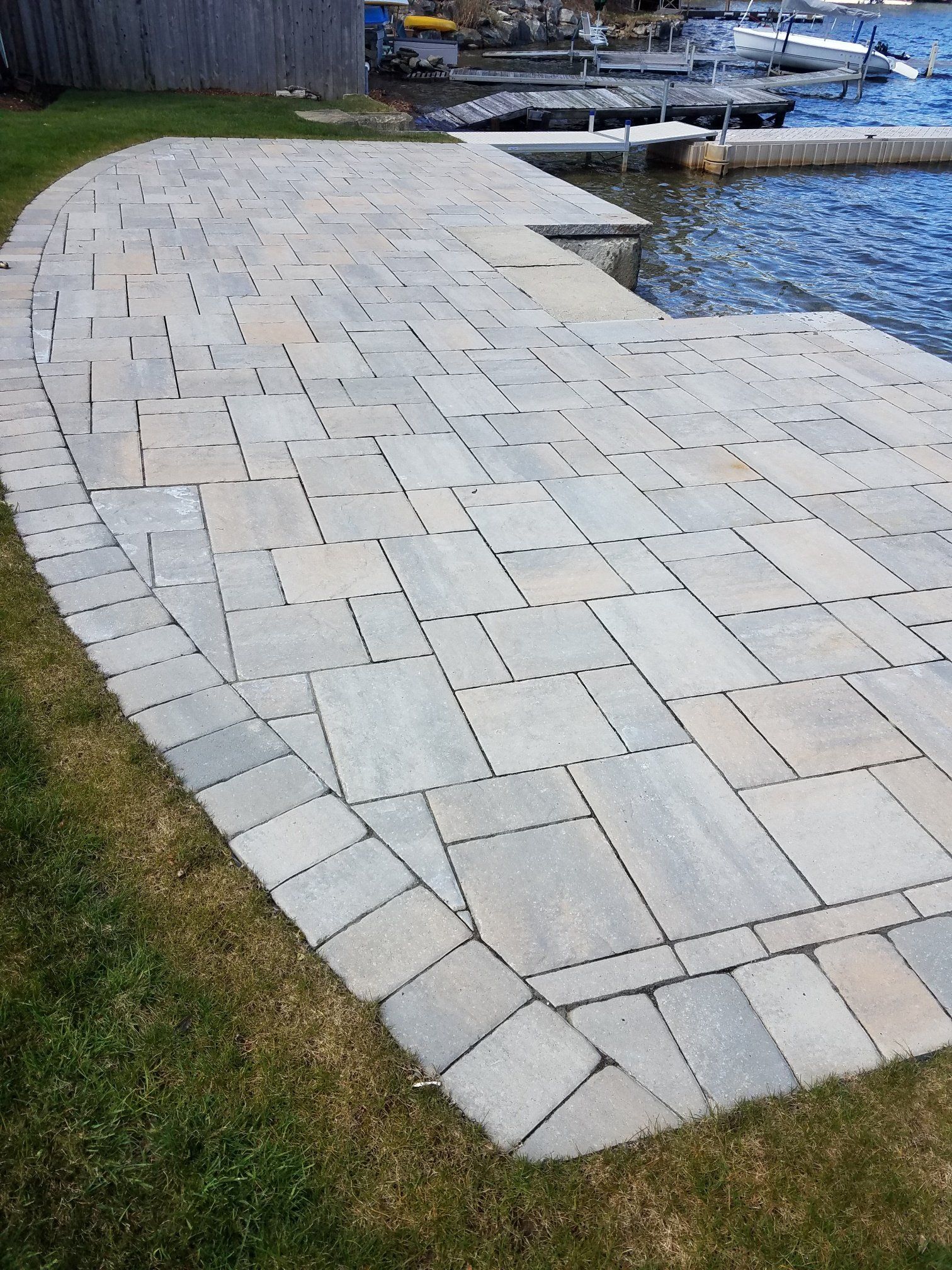 Stone patio beside a lake, leading to a dock with boats in the distance.