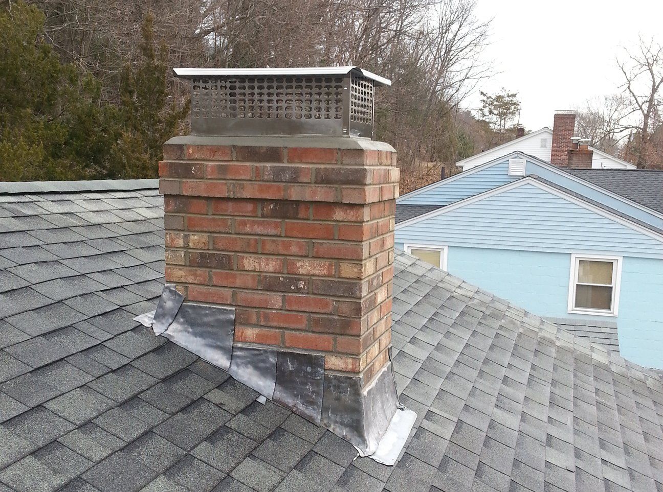 Brick chimney on a gray shingled roof, with a metal cap and flashing. Blue house in the background.