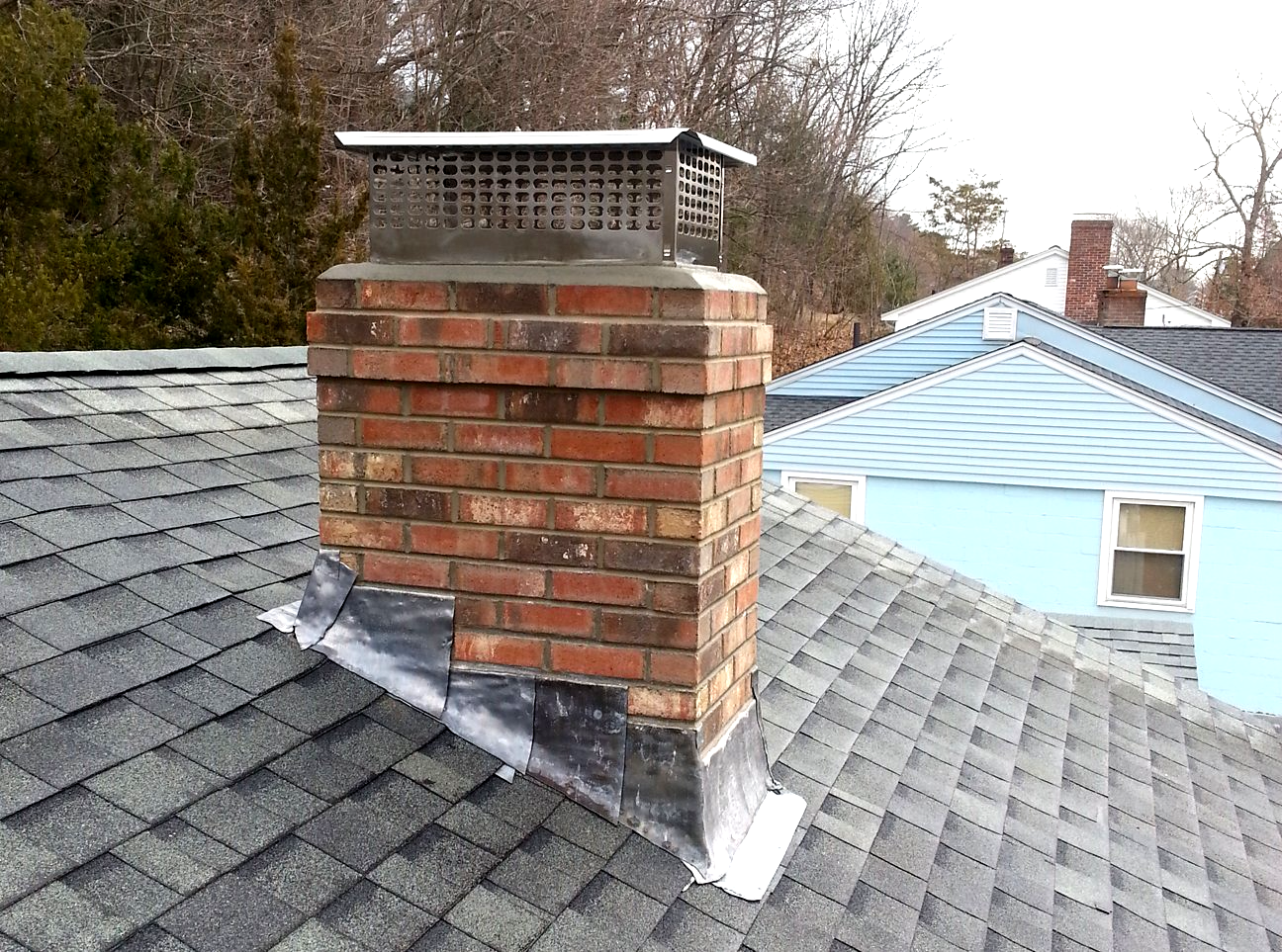 Brick chimney on a shingled roof with a metal cap. Blue house and trees in background.