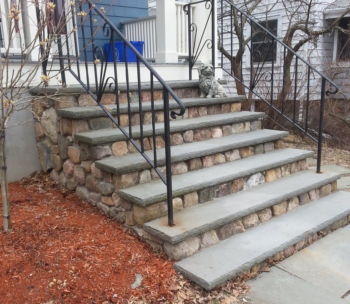 Stone steps with wrought iron railing leading to a house entrance. Red mulch and bare trees surround.