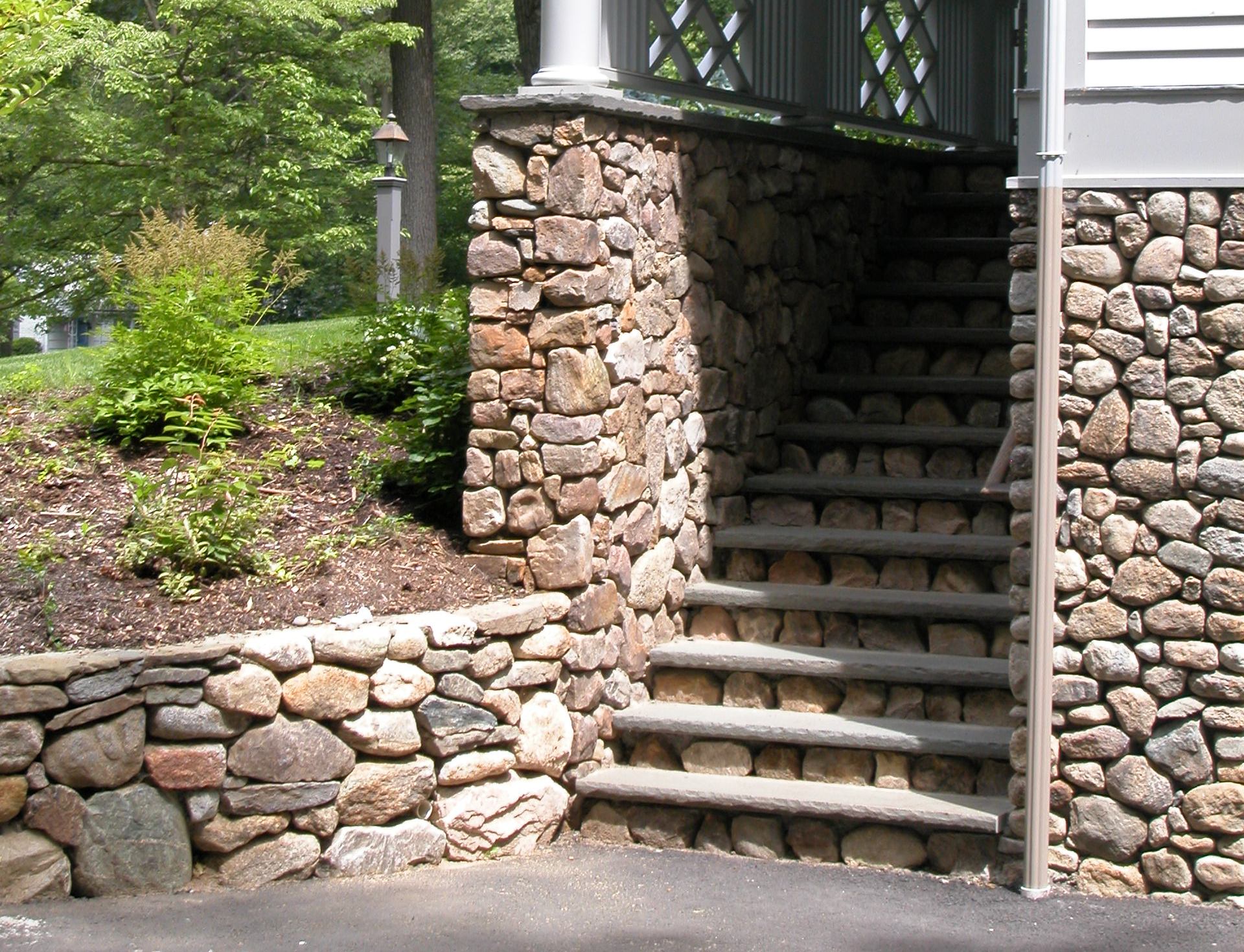 Stone steps leading up to a porch with stone walls, surrounded by greenery.