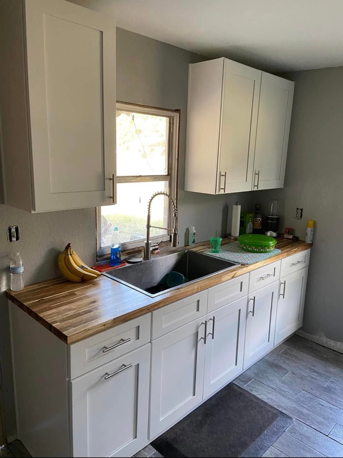 Small kitchen with white cabinets, wooden countertops, double sink, and fruit near a window.